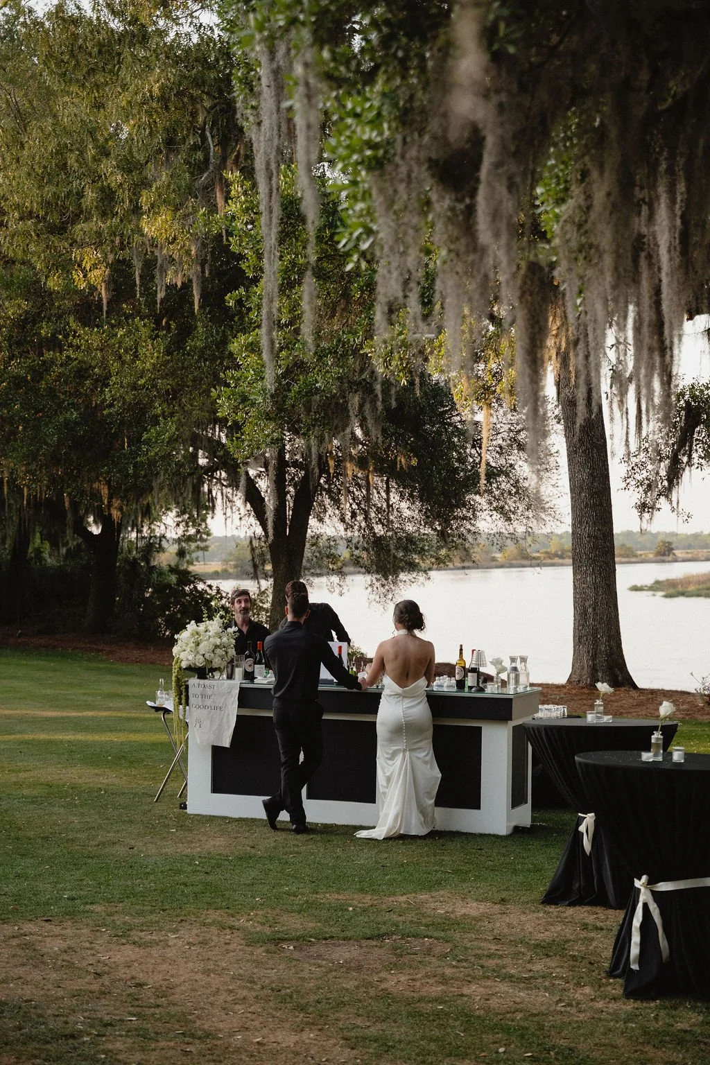 A wedding reception scene outdoors near a lake with large trees draped in moss. A bride in a white gown and a groom are at a black and white bar, talking to a bartender. There are tables with candles and flowers nearby, and the sky appears to be over