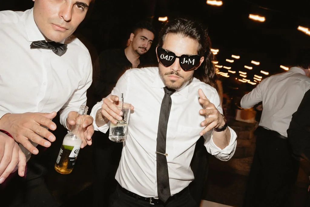 Four men in formal attire, with some wearing white shirts and black ties or bow ties, at a lively event or party at night. The man in the center is wearing sunglasses with white text on the lenses, holding a drink and making a dynamic gesture.