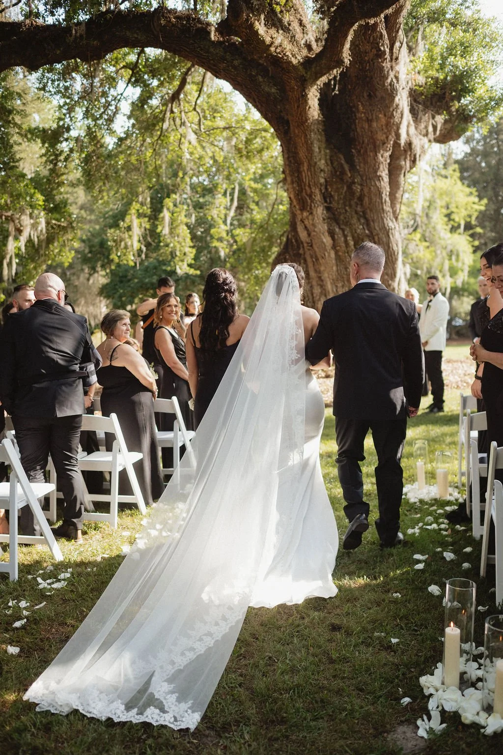 A bride and groom walking down the aisle outdoors during their wedding ceremony, surrounded by guests and large trees with hanging moss.