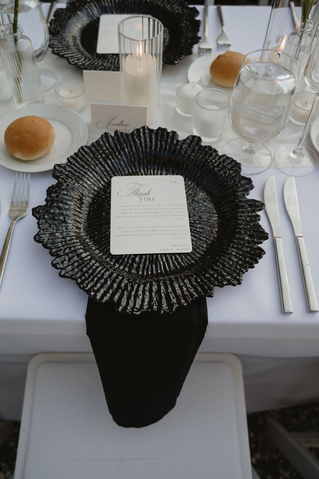 A formal dining table setting with a black textured charger plate, a white thank you note on the plate, a bread roll on a small white plate on the left, clear water glasses, a black candle holder with a lit candle, and utensils including a fork, knif