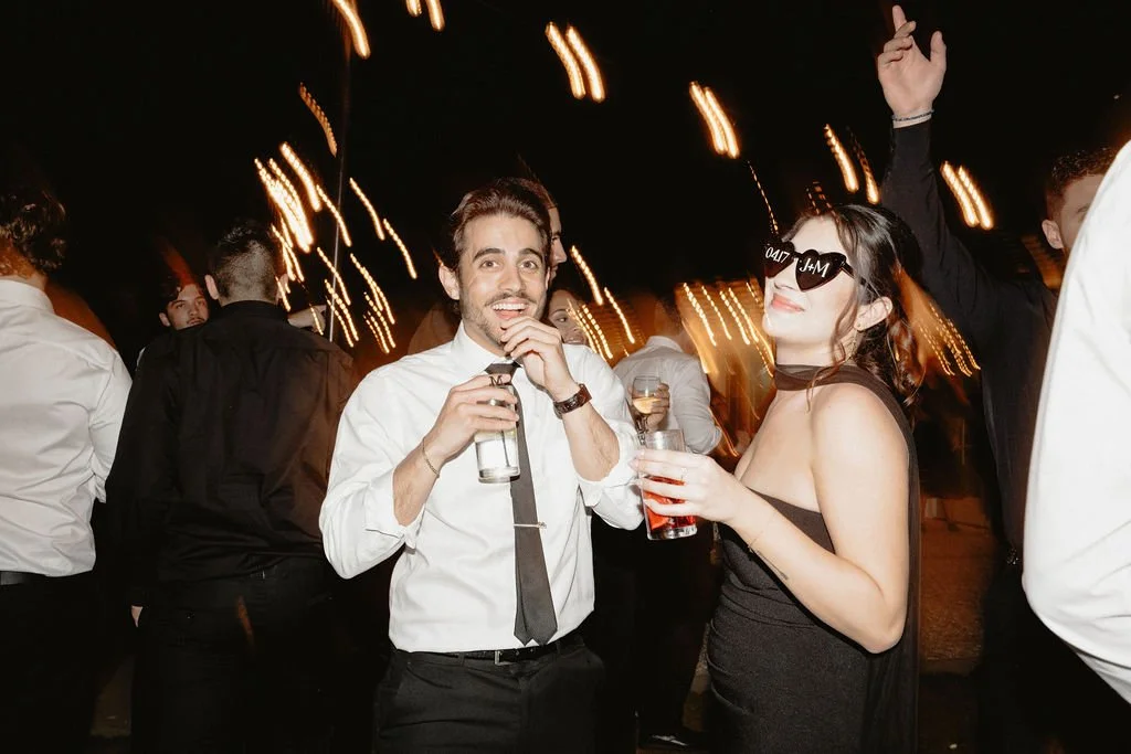 A group of people partying at night, with a woman wearing sunglasses and a woman smiling with a drink in her hand in the foreground.