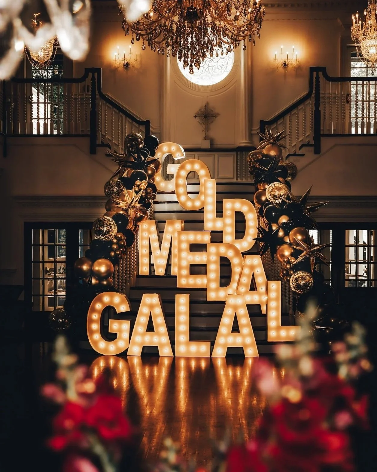 Decorative indoor event space with large illuminated letters spelling "GOLD MEDAL GALA" on stairs, framed by black and gold balloons and star-shaped decorations, under ornate chandeliers and a large circular window in a luxurious setting.