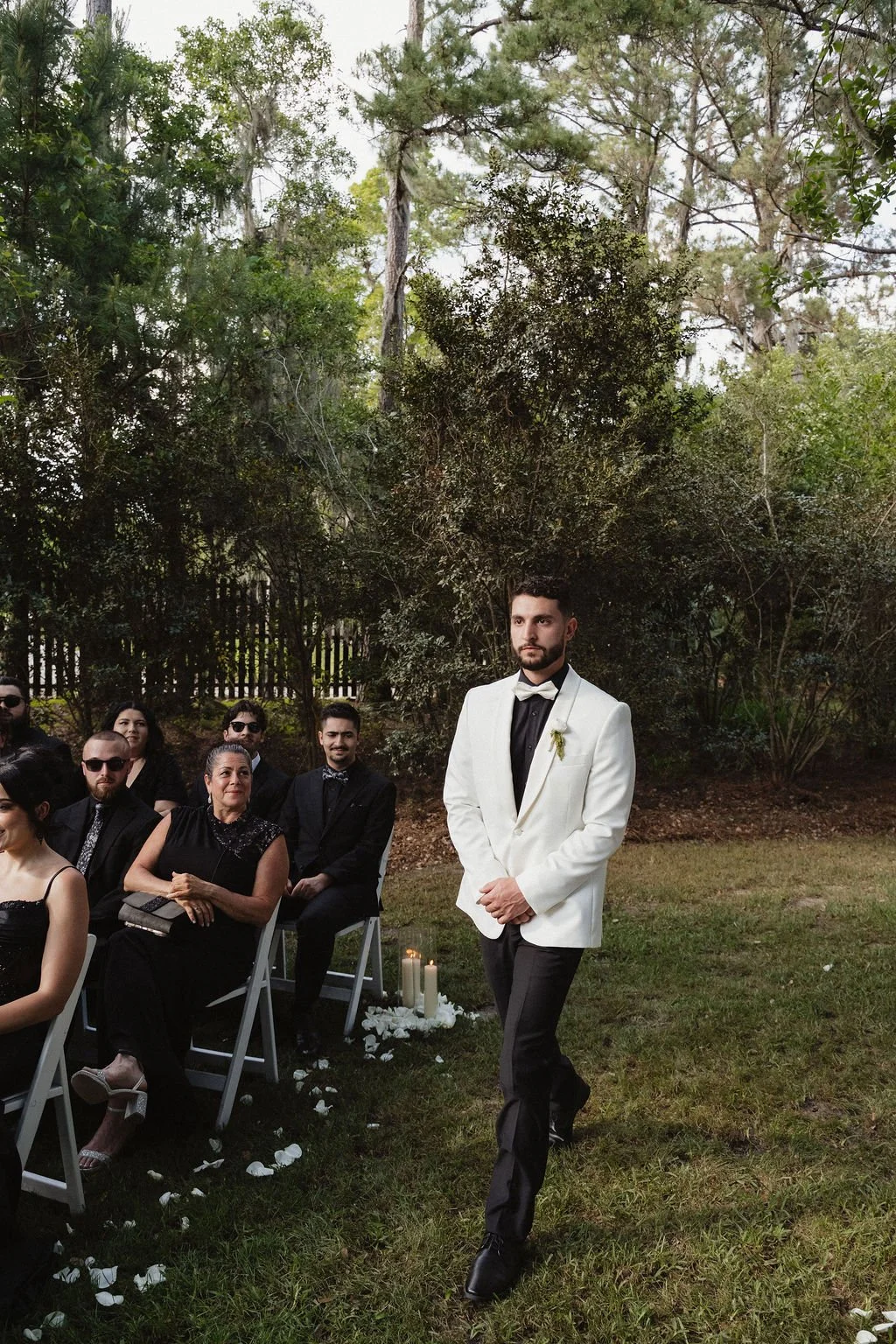 A wedding ceremony outdoors with a groom in a white tuxedo jacket and black pants standing alone, surrounded by seated guests on chairs, in a garden setting with trees and greenery in the background.