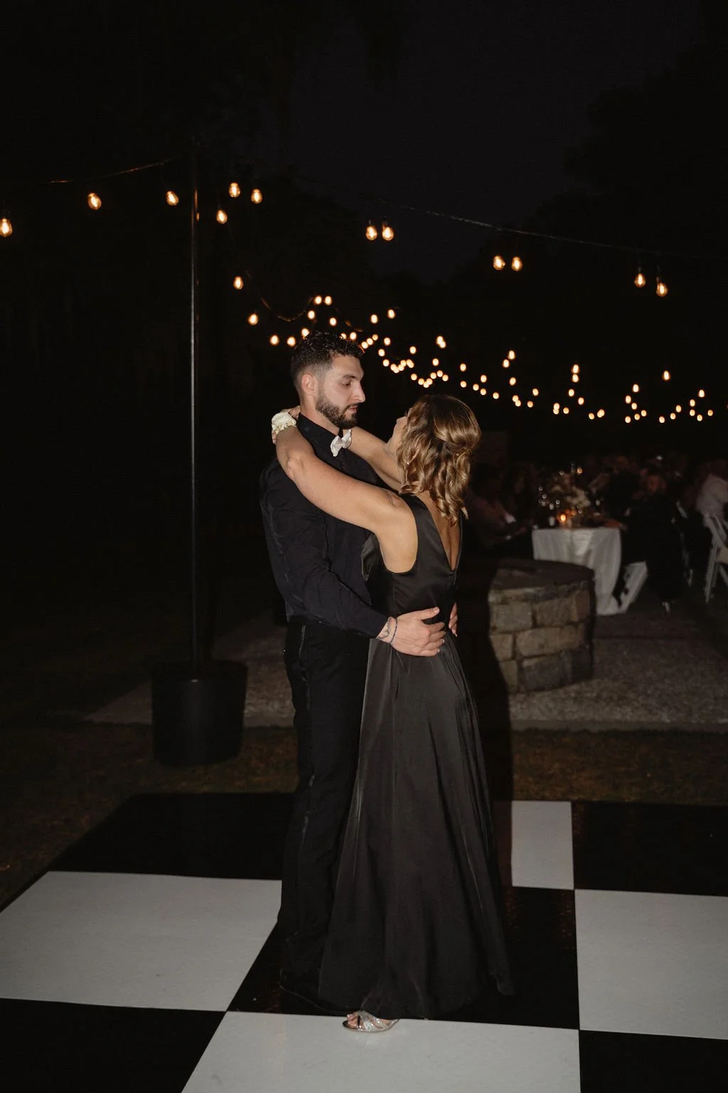 A couple dancing closely on a checkered dance floor at night, under string lights at an outdoor event.