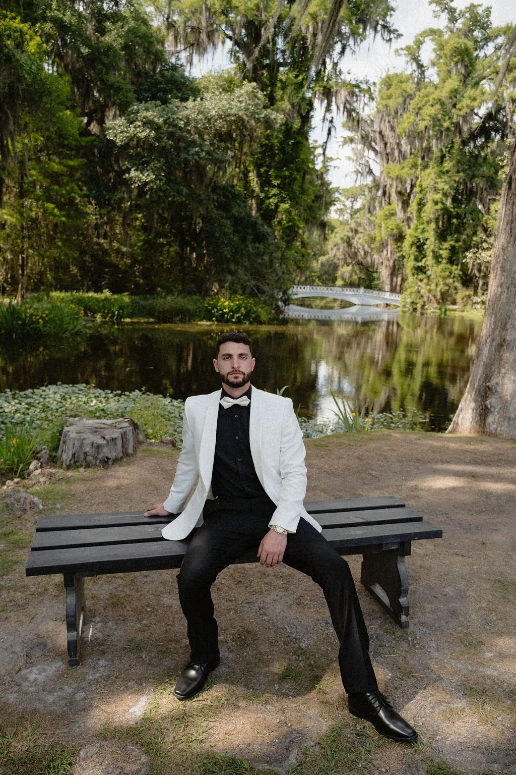 A man in a white tuxedo jacket with a black shirt, black bow tie, and black pants, sitting on a black park bench by a river, with lush green trees and a small white bridge in the background.