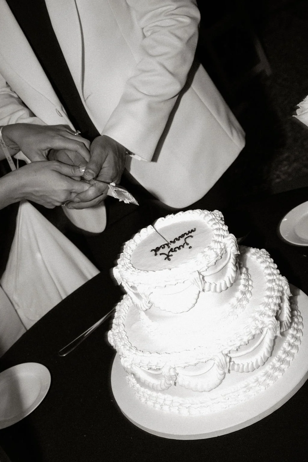 A person in a white suit is cutting a wedding cake with intricate icing details while another person holds their hands. The cake has the words "Just Married" written on the top.