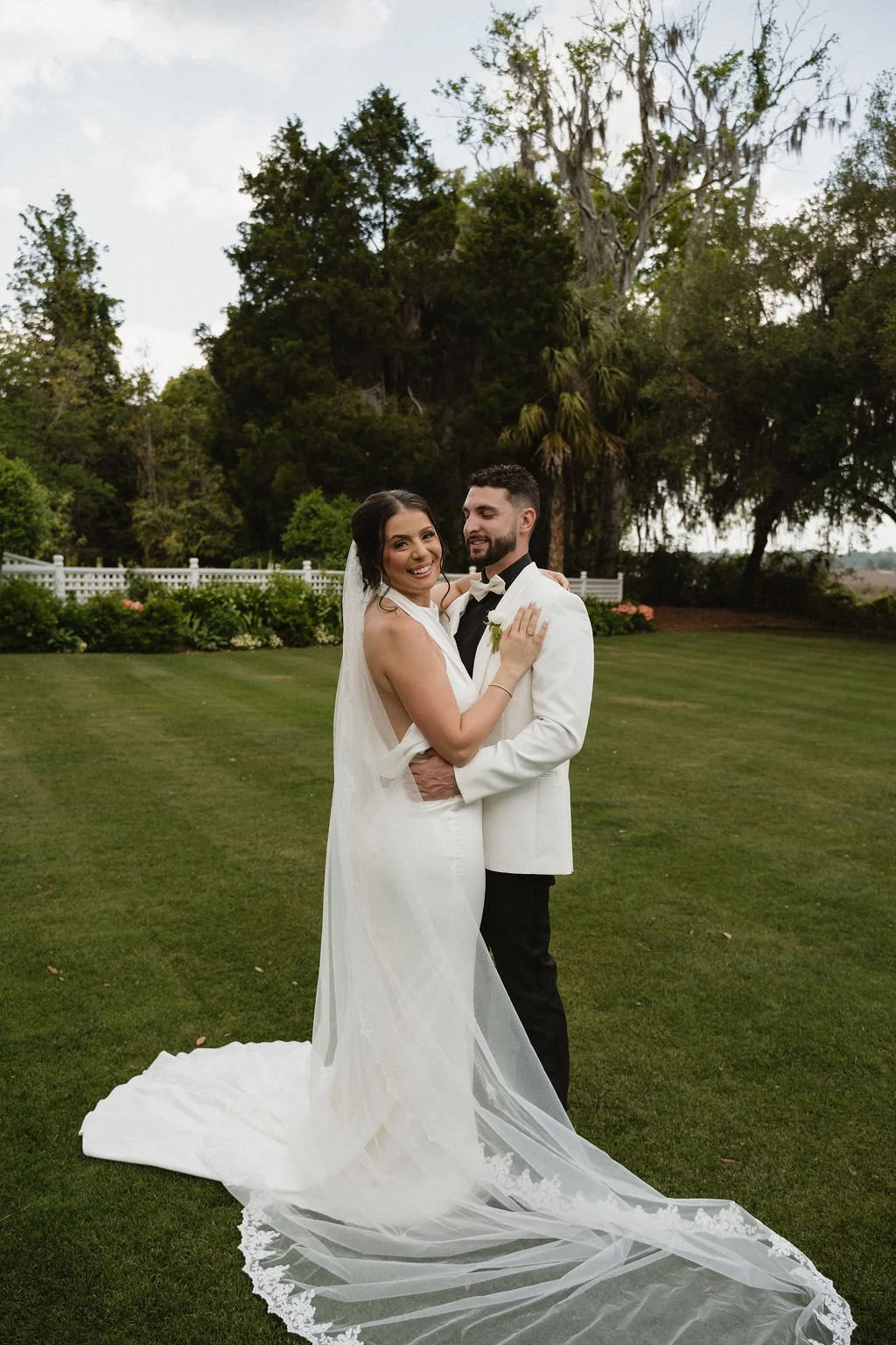 A bride and groom standing on a grassy lawn, smiling and looking at each other, with trees and a white fence in the background during daytime.