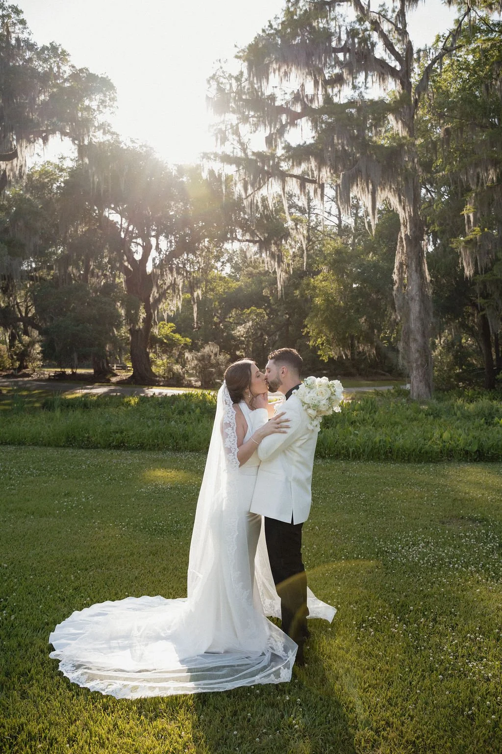 A newlywed couple sharing a kiss outdoors on a grassy area with large trees and Spanish moss, with sunlight shining through the trees.