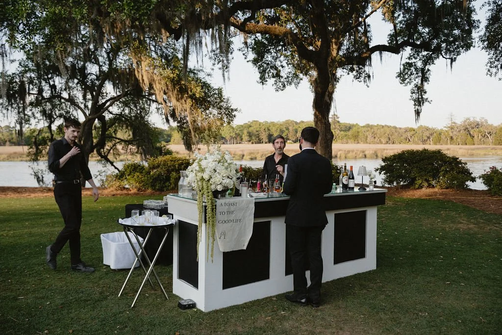 Outdoor gathering with a bar set by a lake, featuring three men in formal attire, two behind the bar, and one in front, with trees and water in the background.