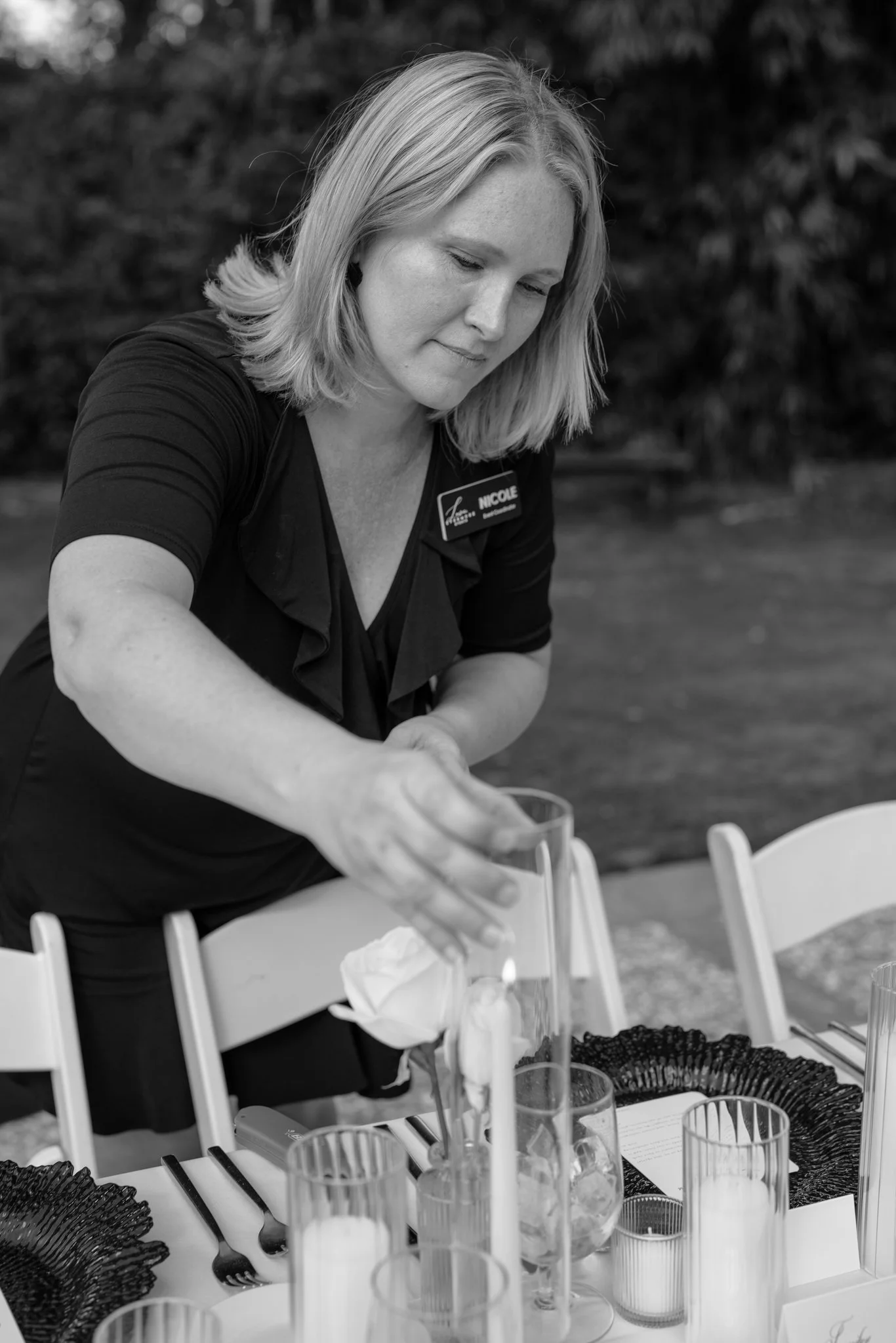 A woman wearing a name tag that says 'Nicole' is preparing drinks at an outdoor table, with various glasses and drink accessories on the table in front of her.