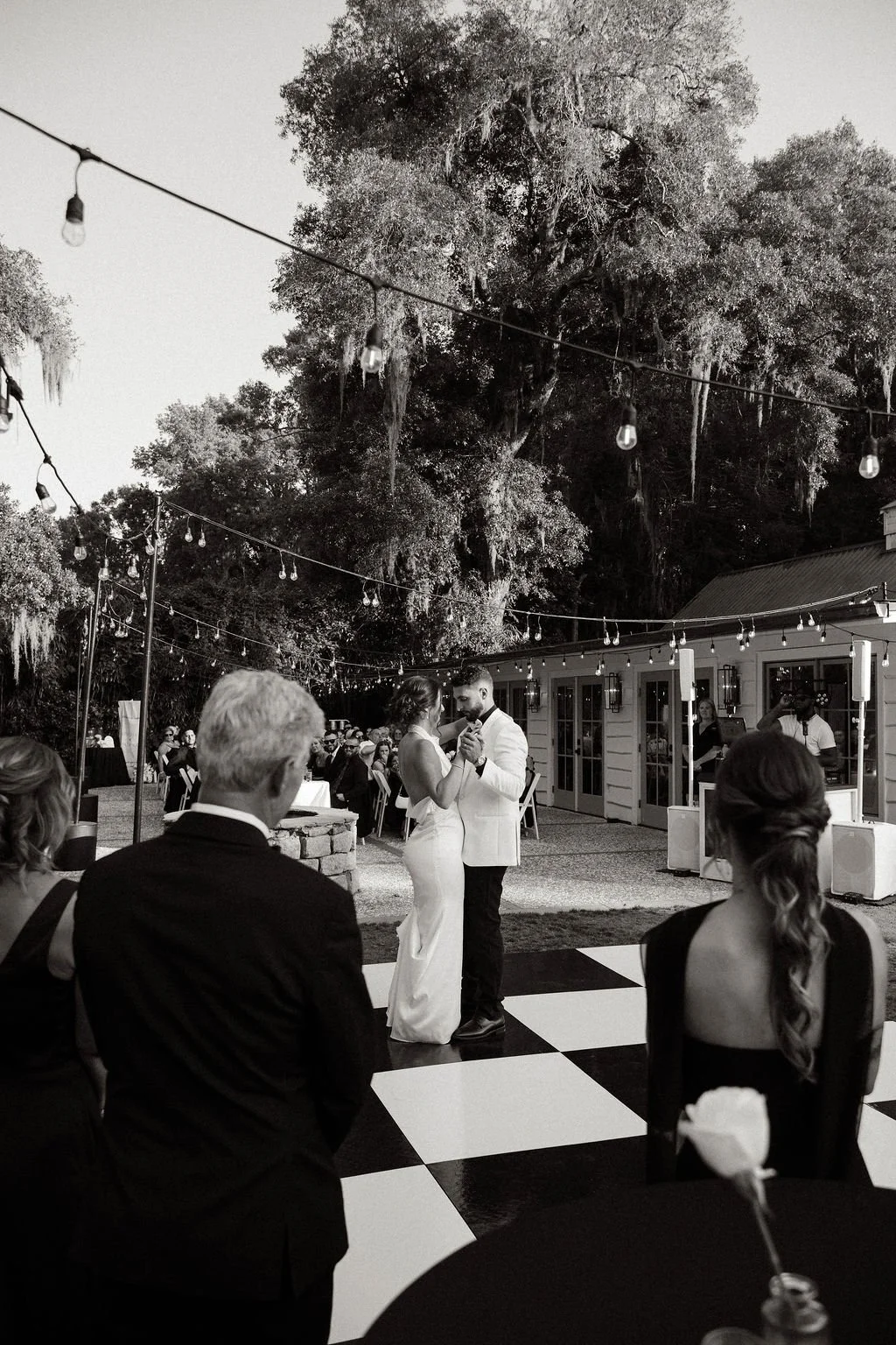 A black-and-white photo of a wedding reception outdoors, with a couple dancing in the center, surrounded by seated guests, string lights overhead, and large trees in the background.