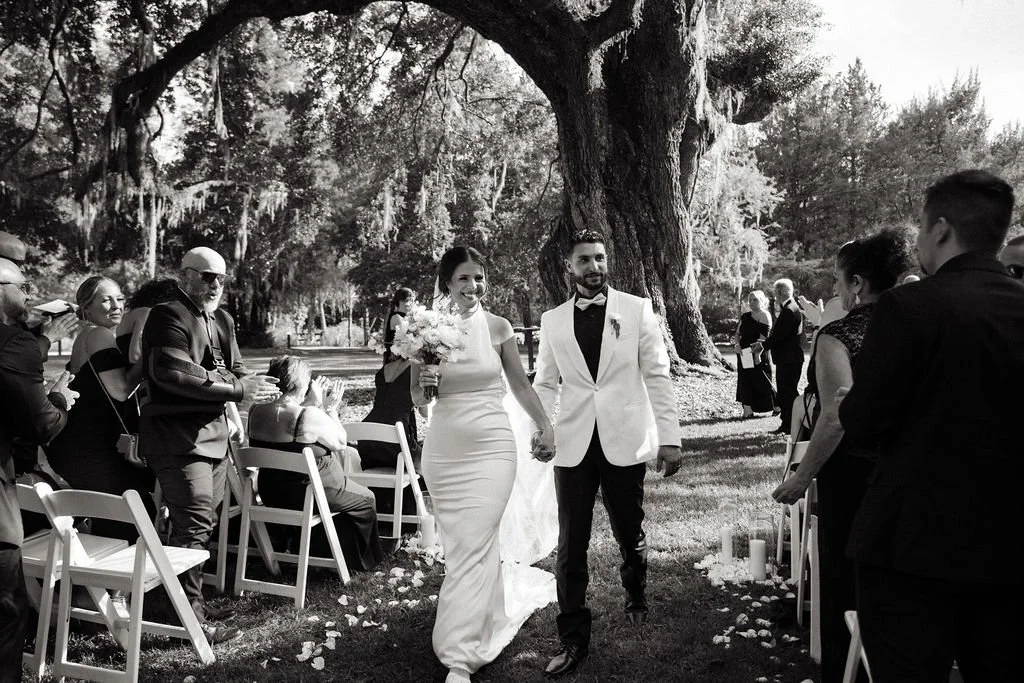 Bride and groom walking hand in hand down the aisle at their outdoor wedding, surrounded by seated guests on either side, with large trees and greenery in the background.