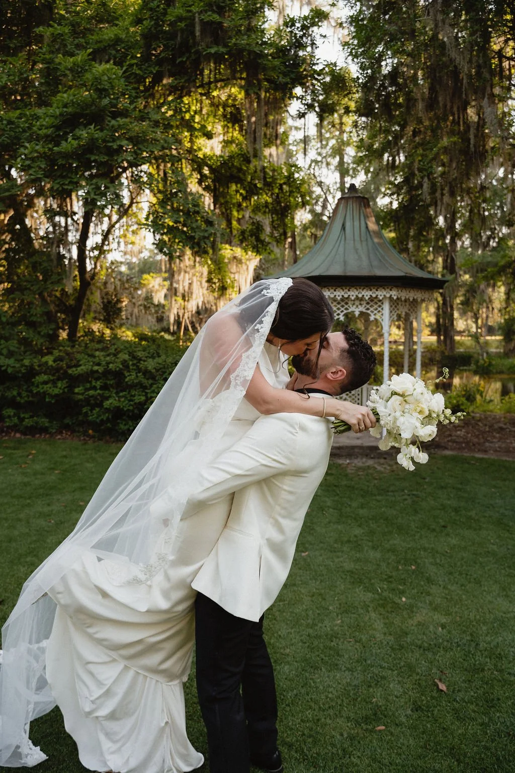 A bride and groom share a kiss outdoors during their wedding, with the groom lifting the bride who is holding a bouquet of white flowers and wearing a white wedding dress and veil, against a backdrop of lush green trees and a decorative gazebo.