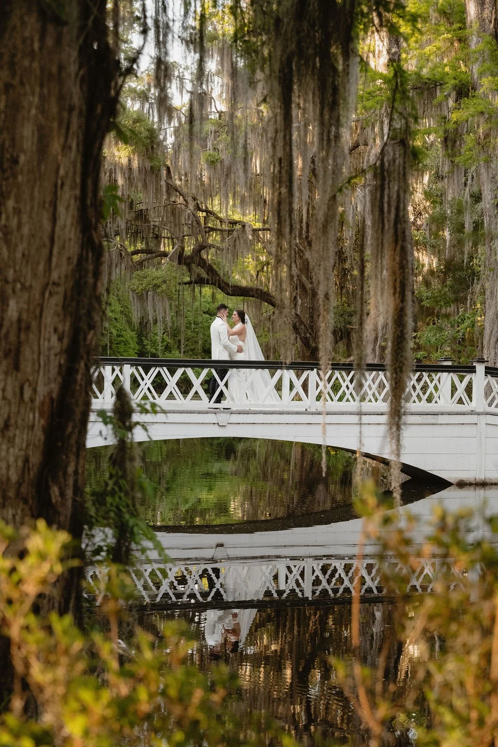A bride and groom dressed in wedding attire standing on a white bridge surrounded by a lush forest with hanging moss and trees.