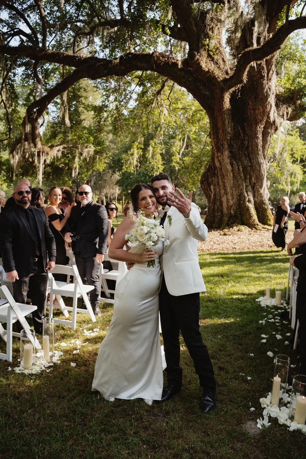 A newlywed couple poses for a photo at an outdoor wedding ceremony under a large tree, surrounded by friends and family.