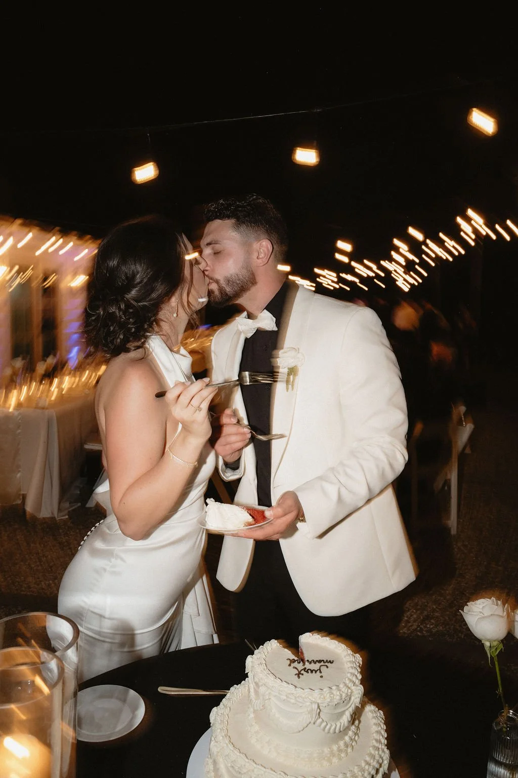 A newlywed couple sharing a kiss at their wedding reception, with a wedding cake in the foreground, evening lighting, and string lights overhead.