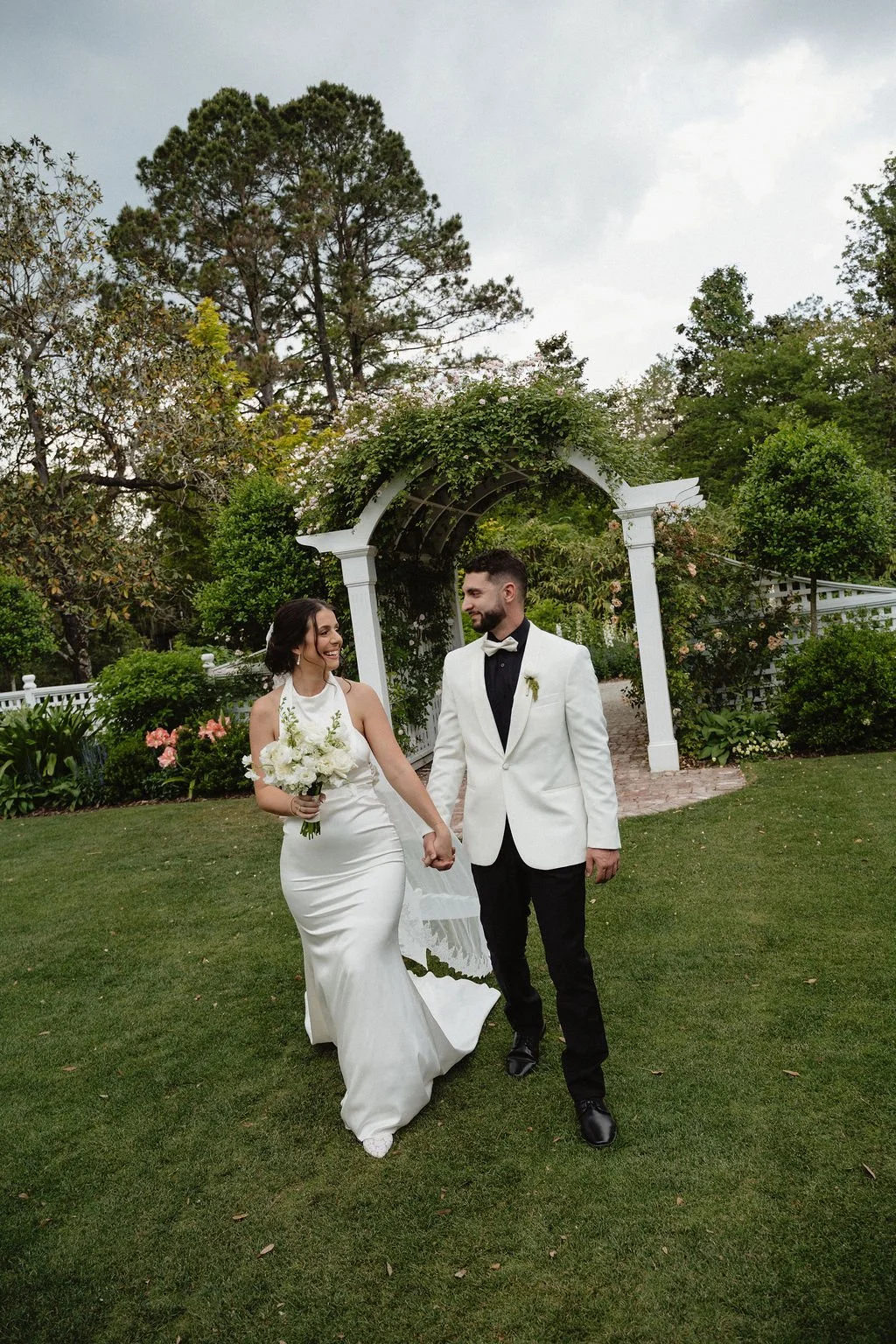 A bride and groom holding hands and walking in a garden during their wedding. The bride wears a white gown and holds a bouquet, and the groom wears a white tuxedo jacket with black pants. They are smiling at each other under a white garden arch, surr