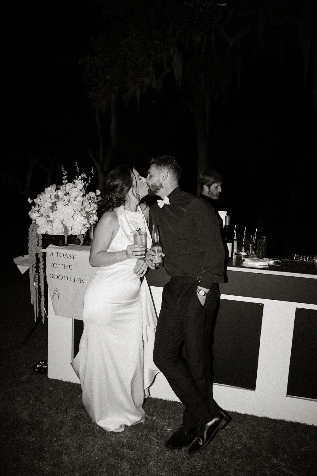 Couple kissing at a wedding reception, holding drinks, with a bar and floral arrangement in the background.