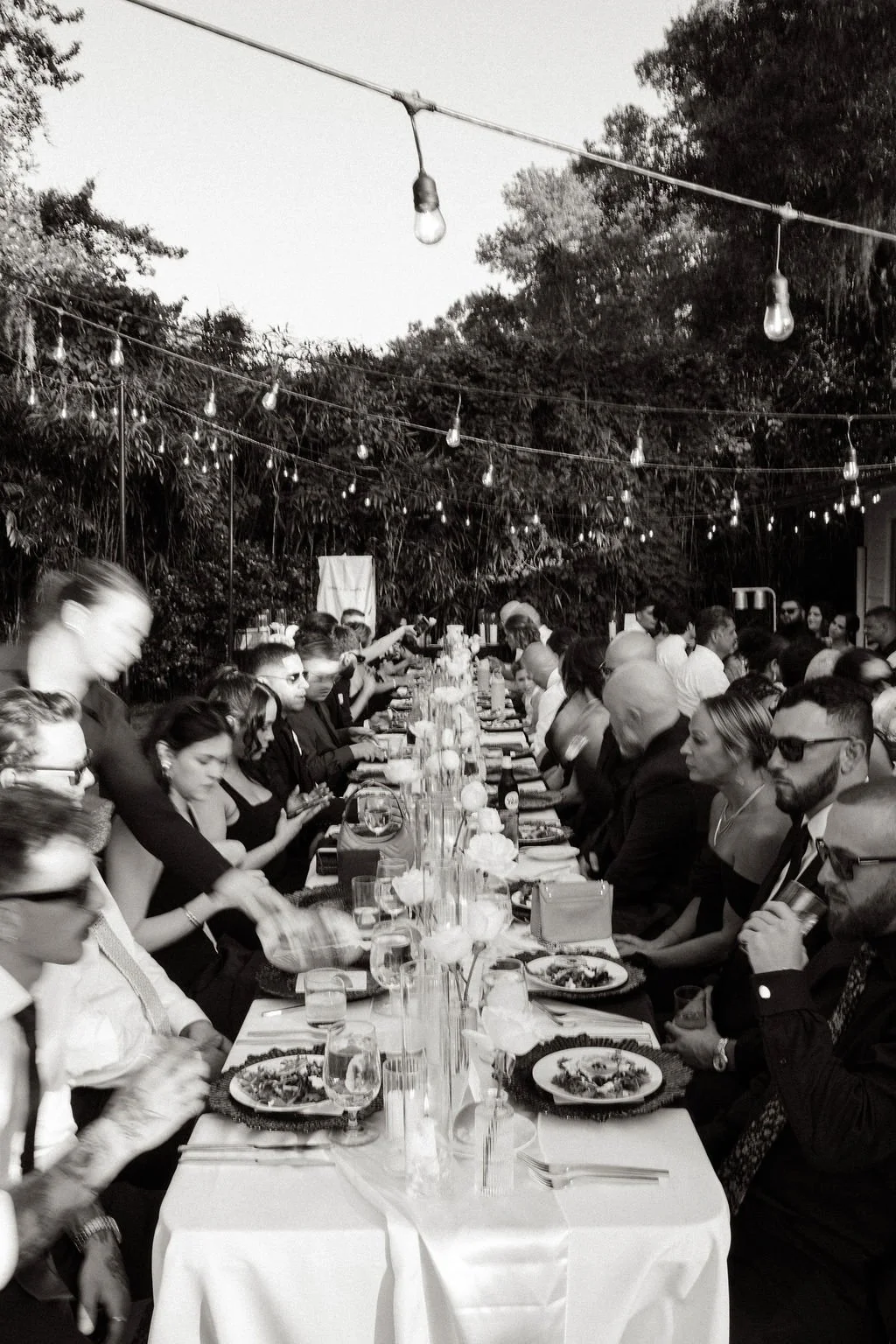 Black and white photo of an outdoor dinner party with many people seated at a long table, decorated with flowers and candles, under string lights, surrounded by trees.