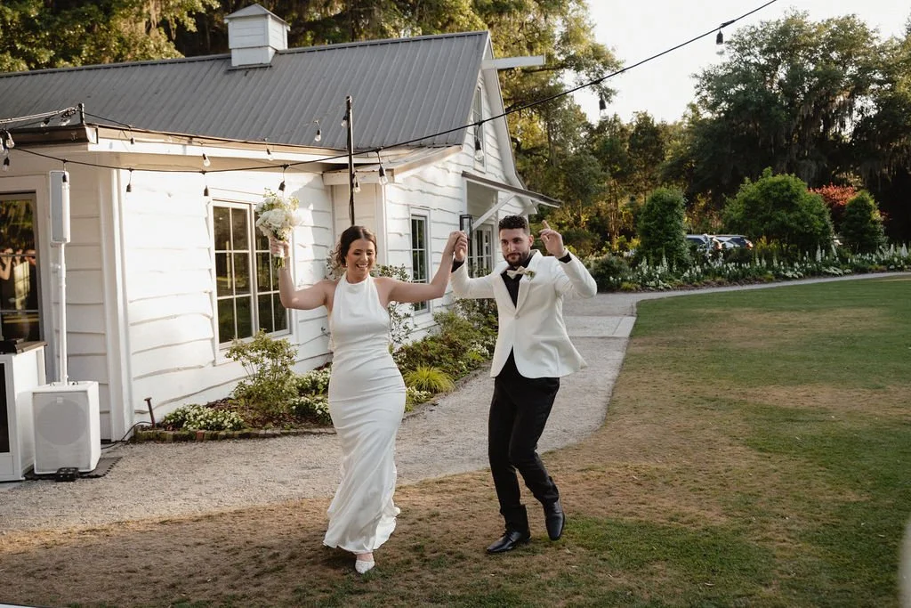 A bride and groom are dancing outside a white house with a metal roof, surrounded by greenery and flowers, during a wedding celebration in the late afternoon or early evening.