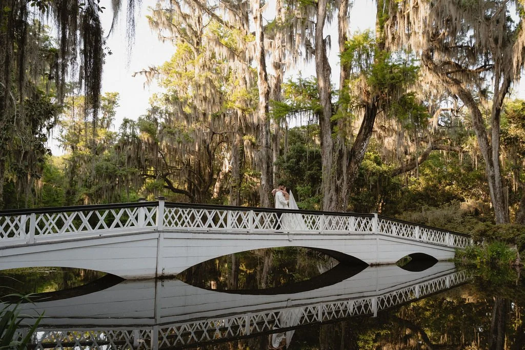 A bride and groom embrace on a white bridge over a pond with Spanish moss hanging from trees in the background.