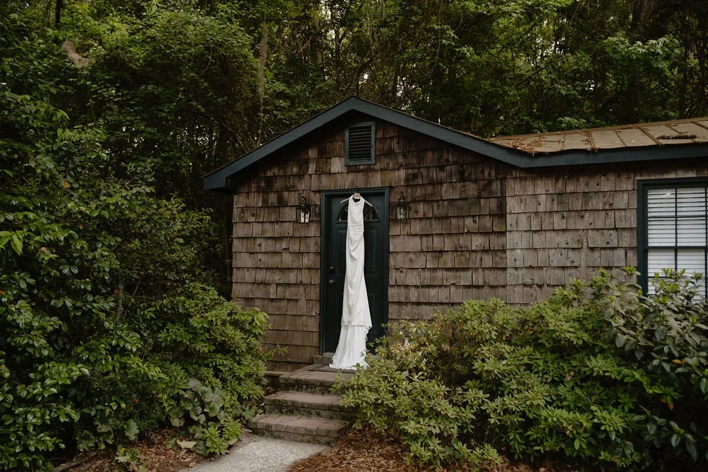 A small wooden house with a dark green front door, surrounded by green bushes and trees. A white cloth or garment hangs on the front door handle, and there are steps leading up to the door.