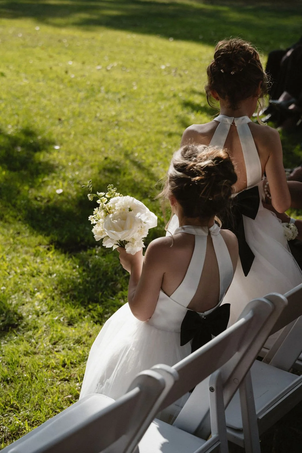 Two young girls in white dresses with black bows, sitting outdoors on white chairs on a grassy area, attending a wedding or event. One girl holds a bouquet of white flowers, and sunlight illuminates the scene.