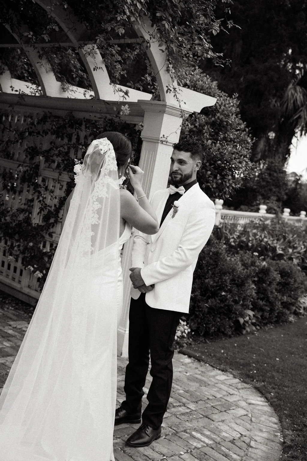 A bride and groom at their wedding, standing outdoors near a garden and gazebo, with the bride adjusting her veil as the groom watches.