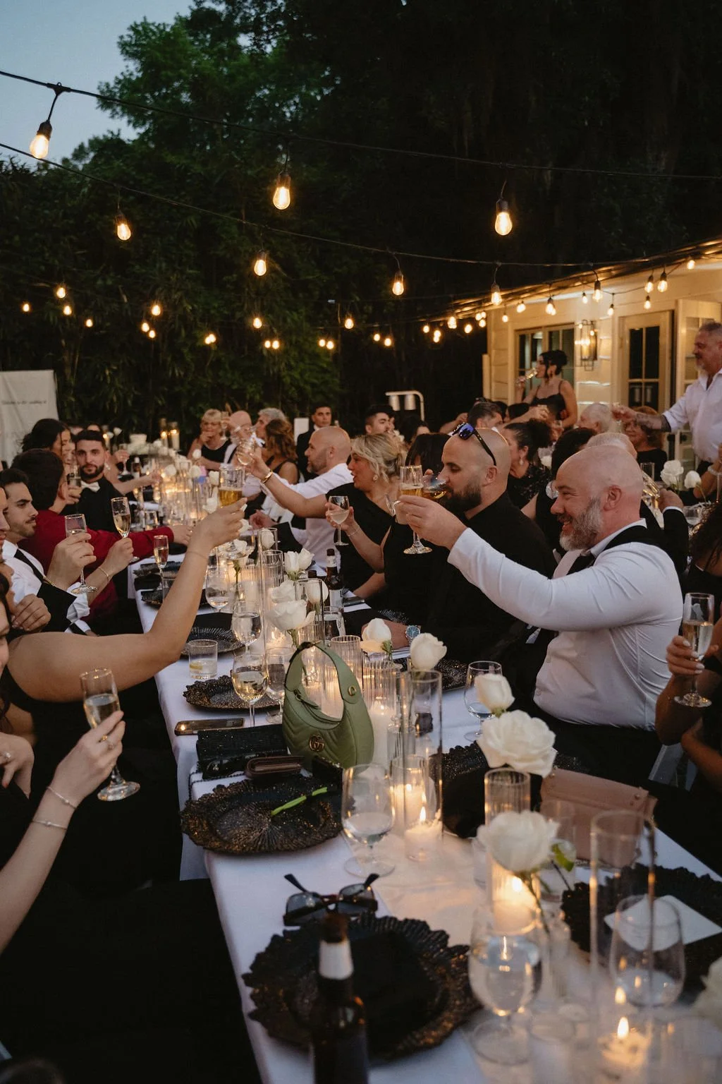 A large outdoor dinner party with many guests sitting at a long table, raising glasses for a toast, decorated with white flowers, candles, and ambient string lights at dusk.
