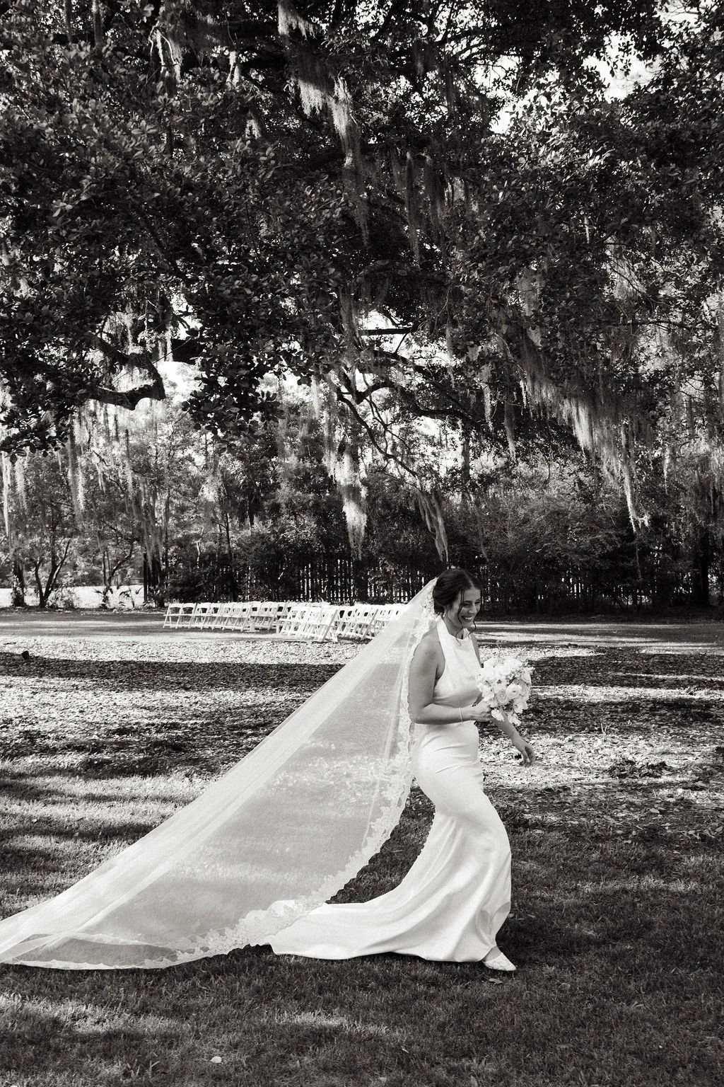 A bride in a sleeveless wedding dress with a long veil walking outdoors on a grassy area, holding a bouquet of flowers, with empty chairs lined up in the background under large trees.