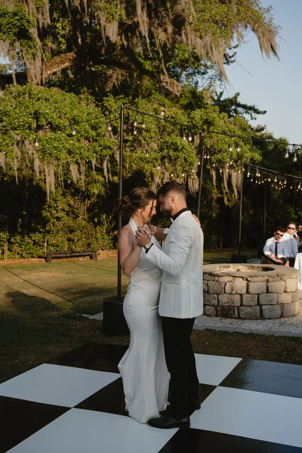 A bride and groom dancing outdoors at sunset, surrounded by string lights, with a small crowd in the background, trees, and a stone fire pit.