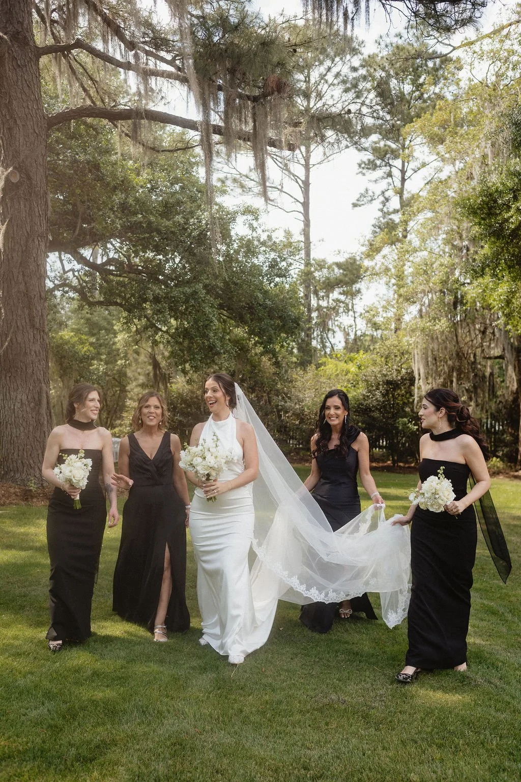 A bride in a white wedding gown and veil walking with four bridesmaids in black dresses holding bouquets of white flowers, outdoors in a lush green park with tall trees.