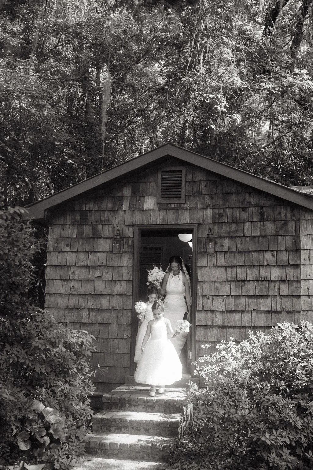 A bride and three flower girls walking out of a rustic wooden chapel surrounded by trees and bushes.