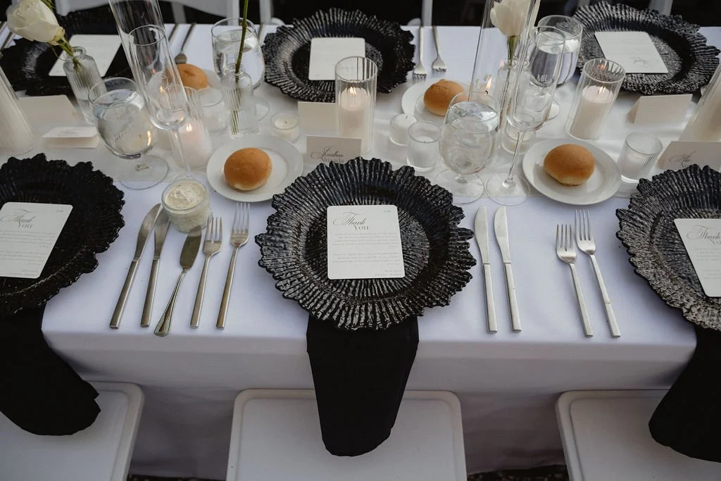 Elegantly set dining table with black textured plates, clear wine glasses, silverware, bread rolls on white plates, candles, and small decorative flowers in glass vases, for a formal event or wedding reception.