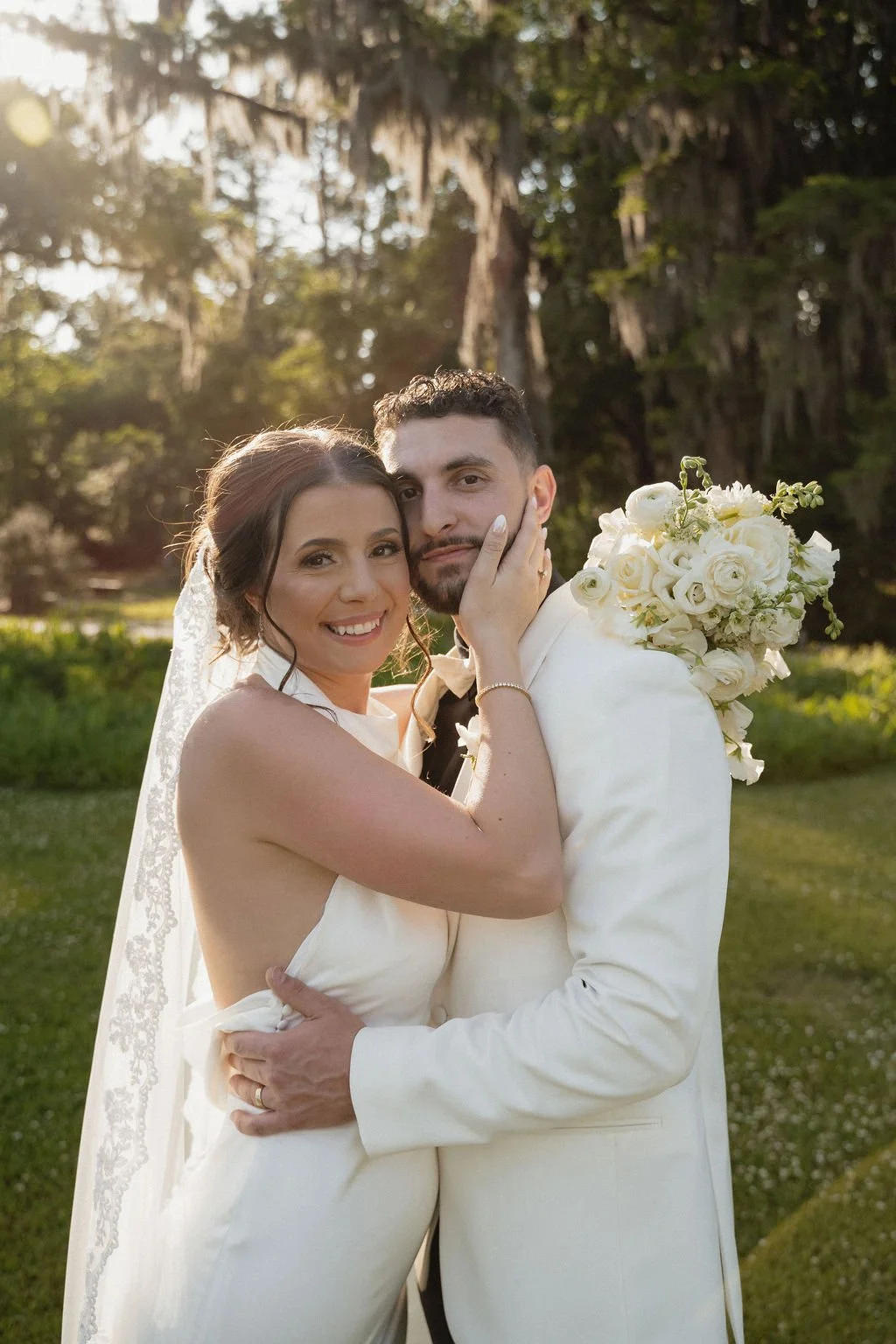 A bride and groom embracing outdoor during sunset in wedding attire, with a bride holding a bouquet of white flowers.