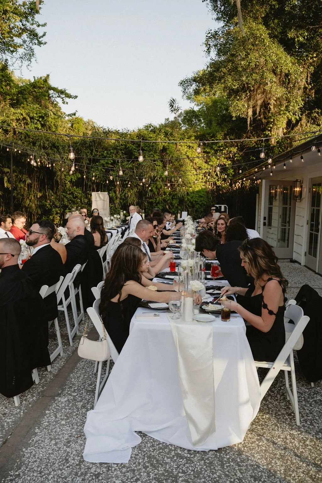 Guests at an outdoor wedding reception seated at long banquet tables on a patio with string lights overhead and lush greenery in the background.