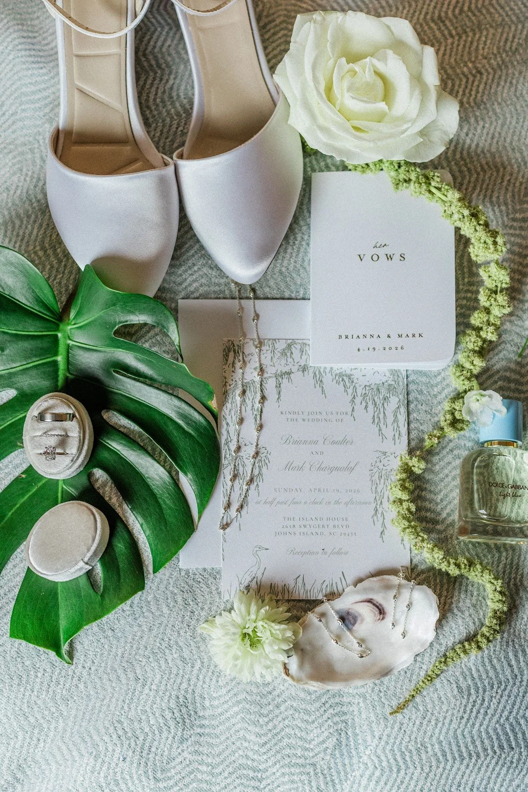Wedding invitation, white bridal shoes, white rose, jewelry, ring box with rings, large green leaf, flower, perfume, and other decorative items on a textured surface.