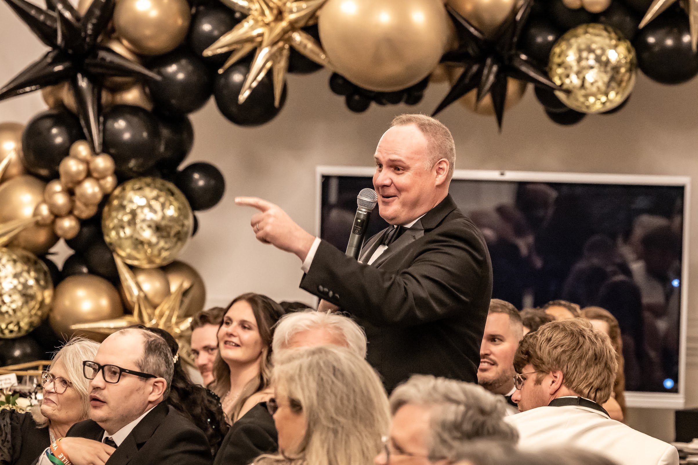 A man in a black tuxedo speaking into a microphone at a formal event with decorated balloons and seated audience members watching him.