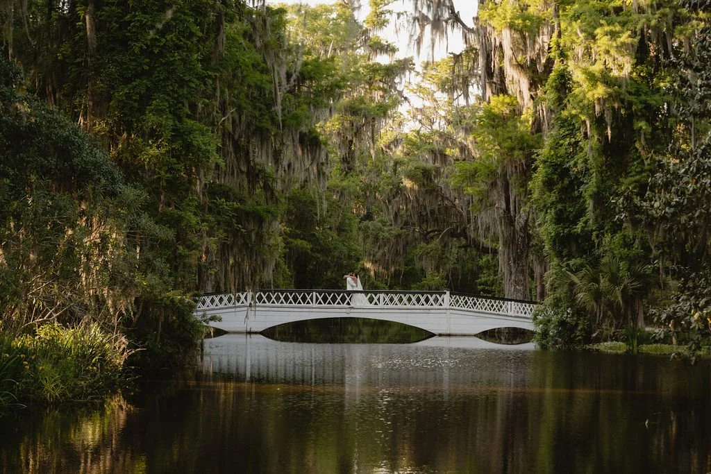 A white arched bridge over a calm river in a lush forest with hanging moss and tall trees.