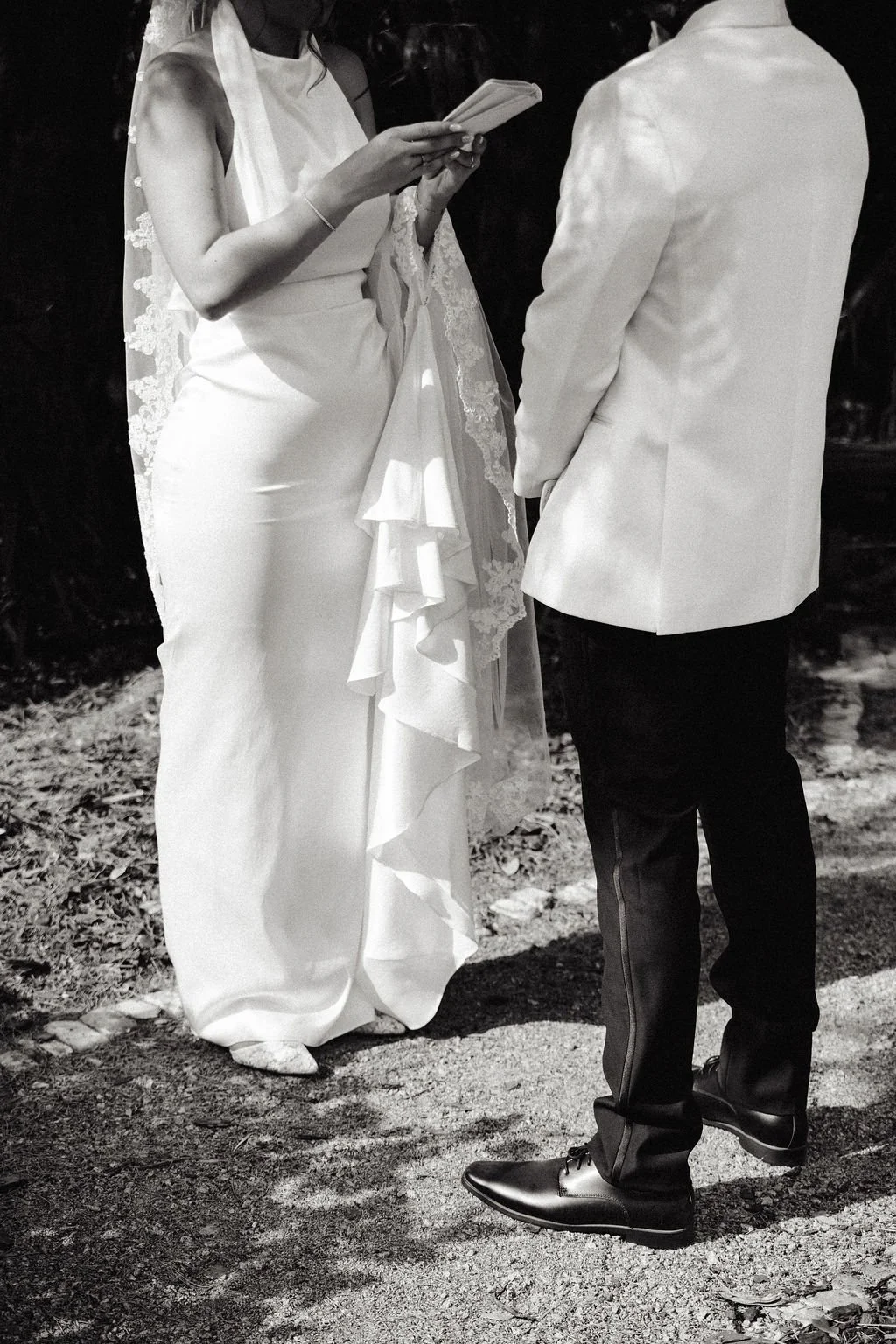 A woman in a white wedding dress holding a book or note, standing in front of a man in a white tuxedo jacket and dark pants, outdoors with a natural background.
