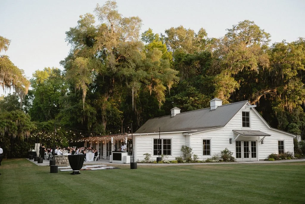 An outdoor wedding reception with guests seated at tables, string lights overhead, and a white house-style building in the background surrounded by trees.