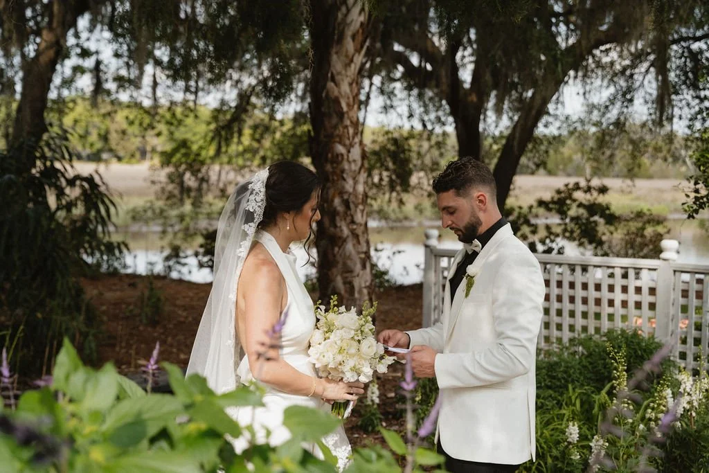 A bride and groom standing outdoors during a wedding ceremony. The bride holds a bouquet of white flowers, and the groom reads vows or a speech. They are in formal attire, with the bride in a white wedding dress and veil, and the groom in a white tux