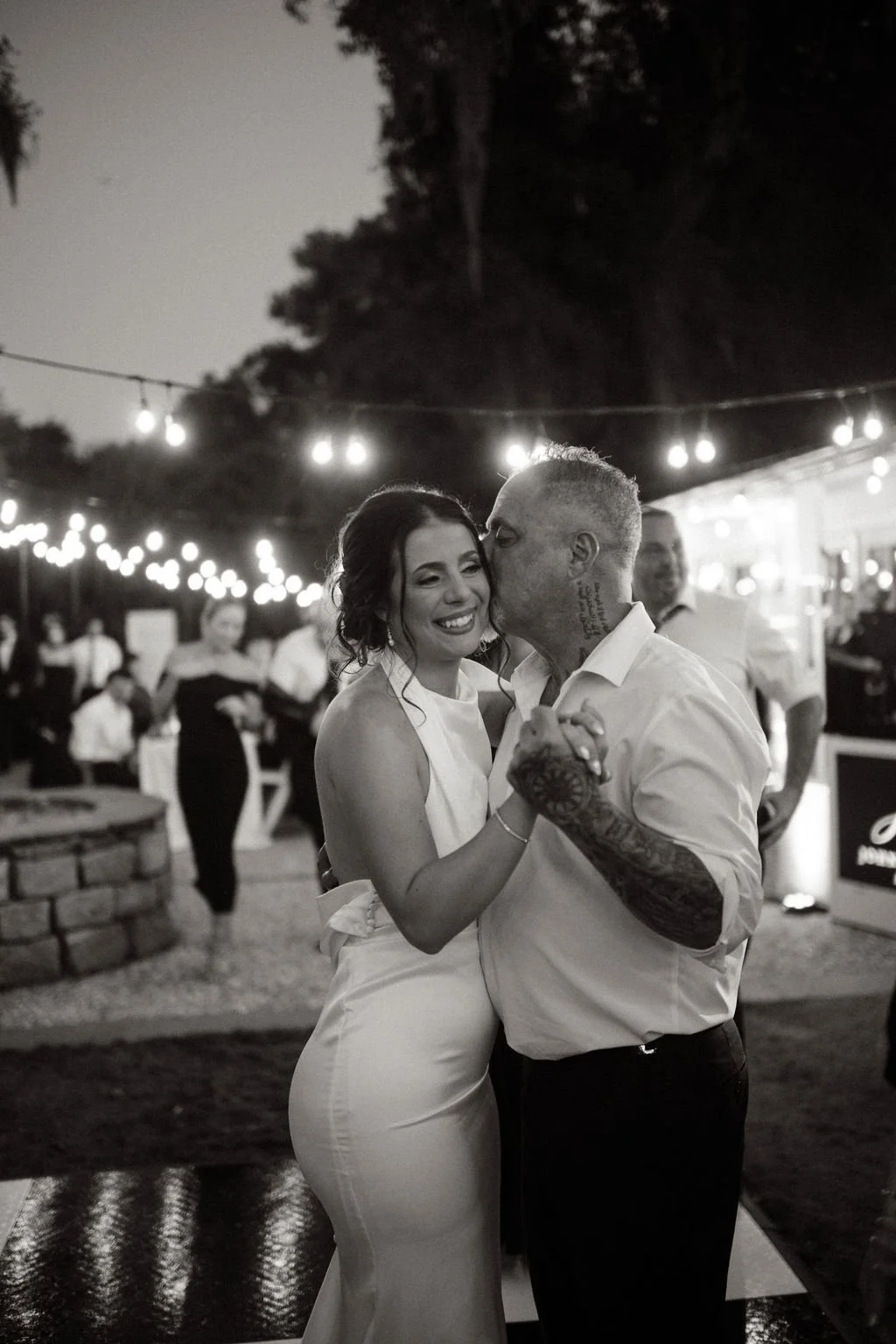 A bride and groom dancing at their wedding reception, smiling, with string lights hanging overhead, in an outdoor setting at night.