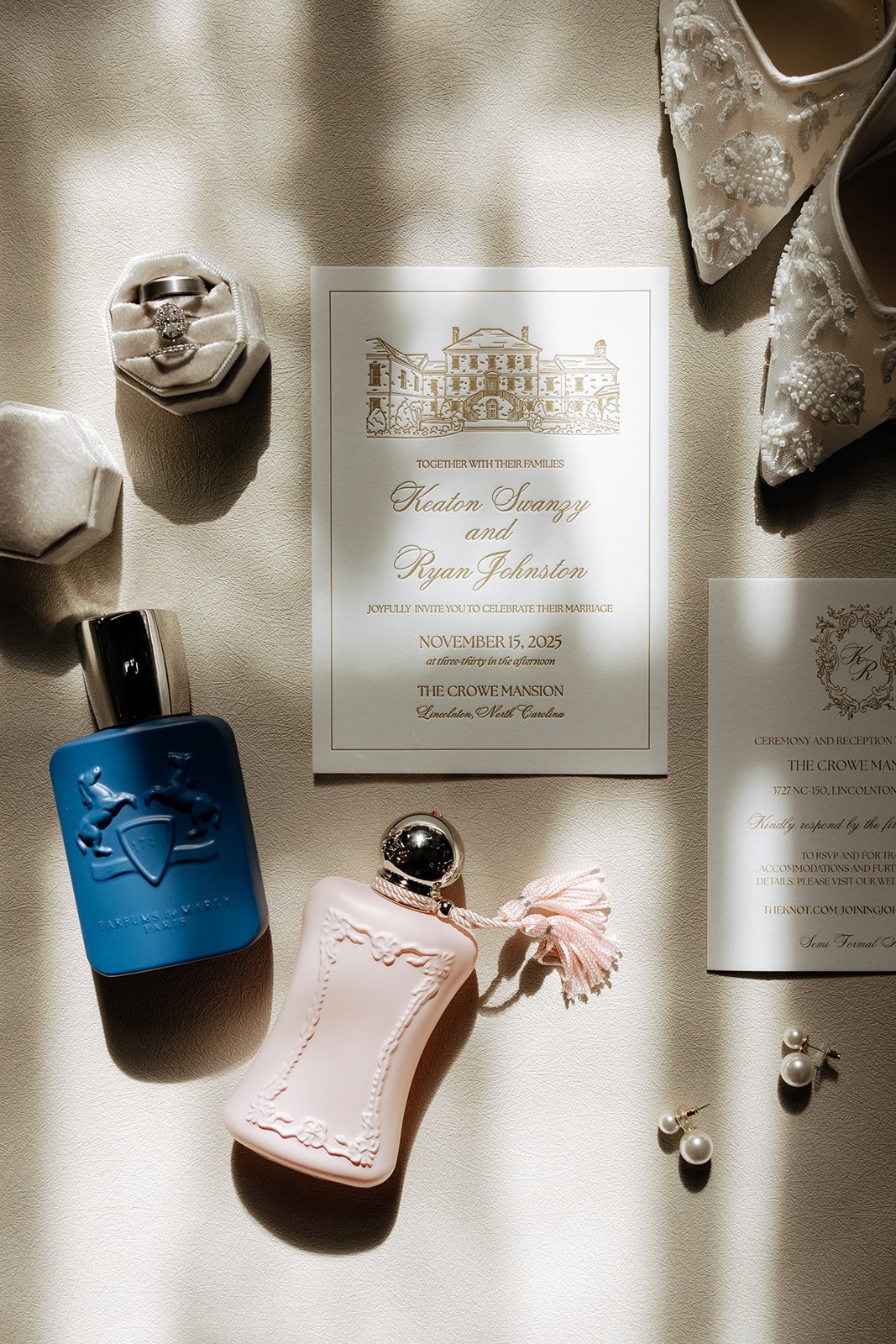 Wedding invitation, perfume bottles, jewelry, and floral shoes on a white fabric surface.