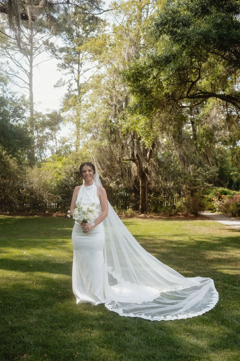 A smiling bride in a white wedding gown holding a bouquet of white flowers, standing on a grassy area under trees with sunlight filtering through