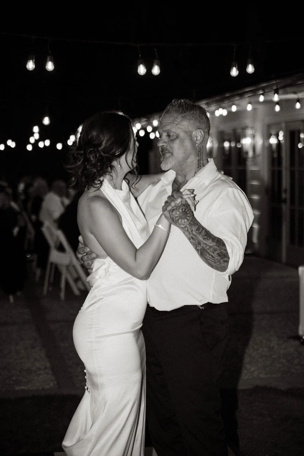 A couple dancing at a nighttime wedding reception with string lights overhead, the woman in a white gown and the man in a white shirt with tattoos on his arms.