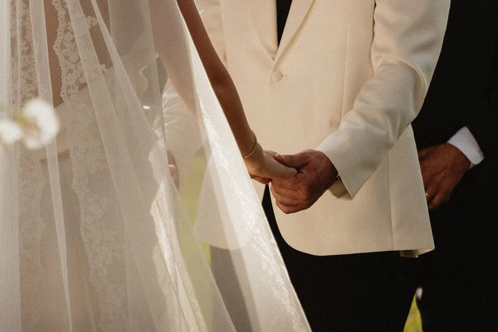 A couple holding hands during a wedding ceremony, with the bride wearing a lace wedding dress and veil, and the groom in a white tuxedo jacket.