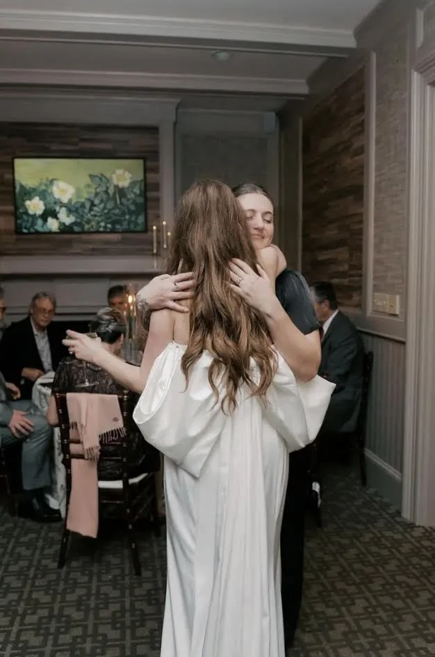 Two women hugging at a celebration, with guests sitting at a table in the background, indoors with wood-paneled walls and a painting of white flowers.