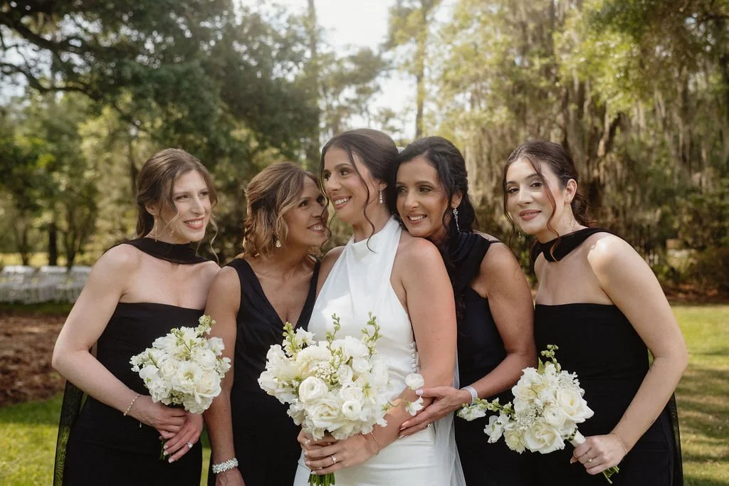A bride in a white dress standing with four women in black dresses outdoors, holding white bouquets, surrounded by trees and greenery.