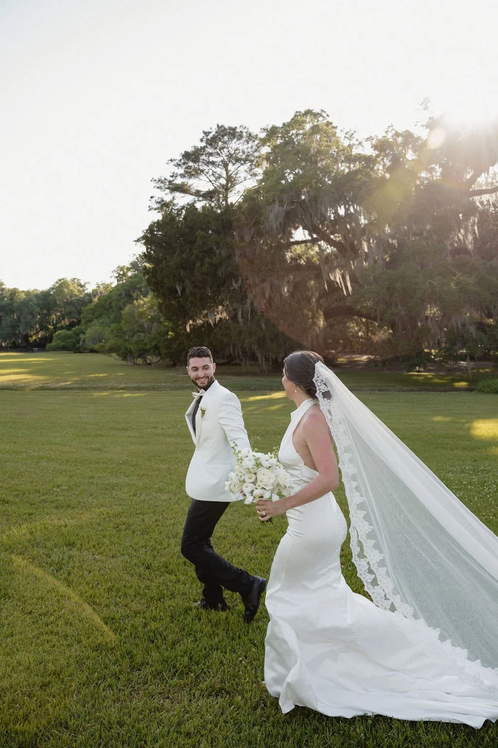 A bride and groom holding hands and walking on a grassy field during sunset, with trees in the background.